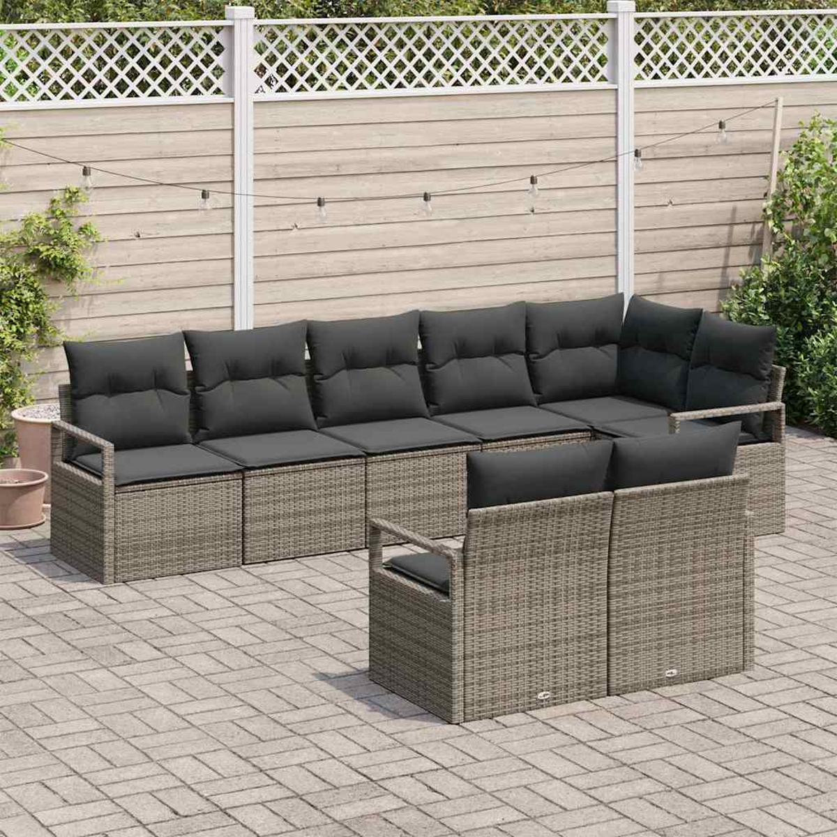 Garten Sitzgruppe auf einer Terrasse. Graue Korbmöbel mit dunklen Kissen. Im Hintergrund ein Zaun und Lichterketten.
