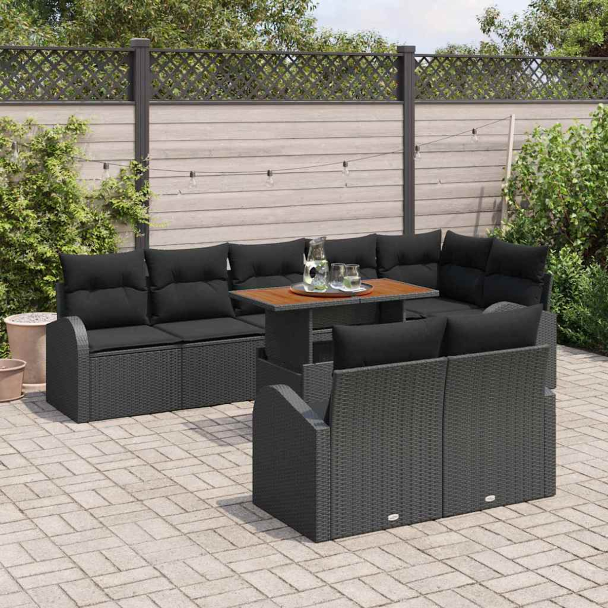 Garten-Sitzgruppe mit schwarzen Kissen und einem Holztisch. Auf dem Tisch stehen eine Kanne und Gläser. Die Gruppe steht auf einer Steinterrasse.