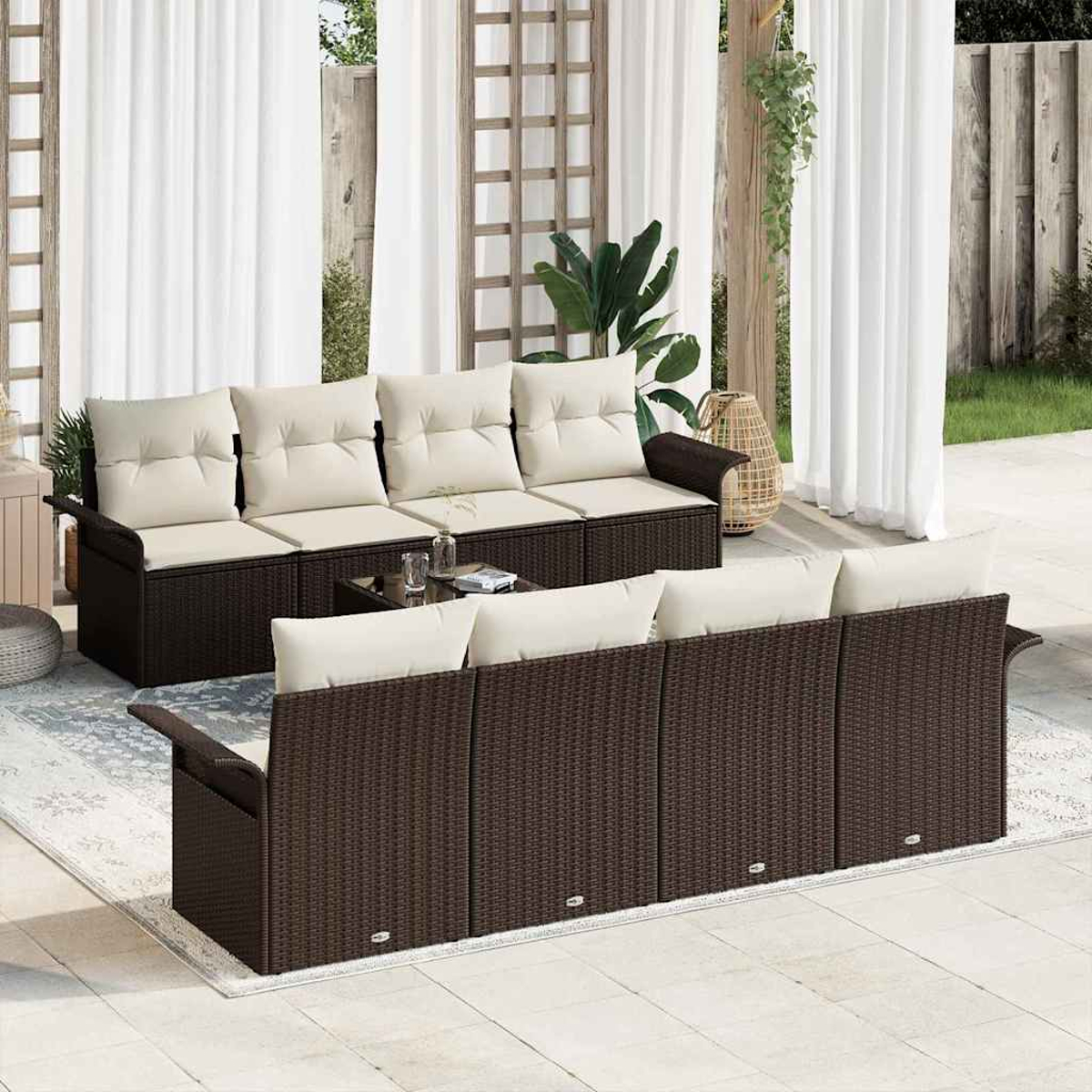 Conjunto exterior con sofás de mimbre marrón oscuro, cojines crema, mesa de cristal y cortinas blancas. Plantas y jardín en el fondo.