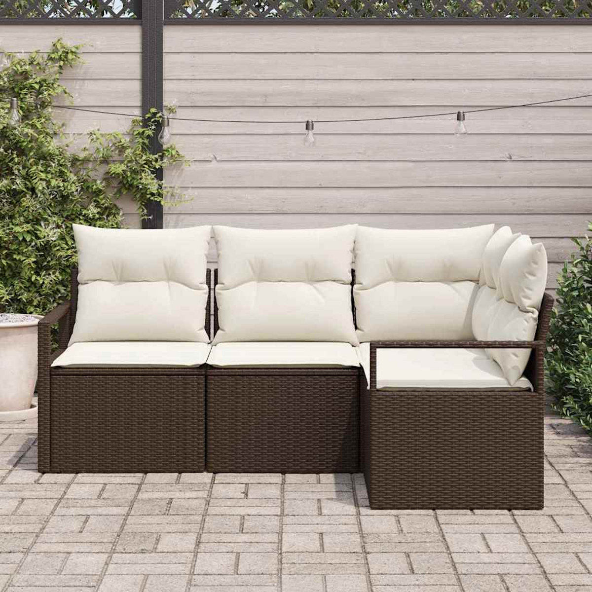 Sofá exterior de mimbre marrón con cojines blancos en un patio pavimentado. Una valla de madera y vegetación en el fondo.
