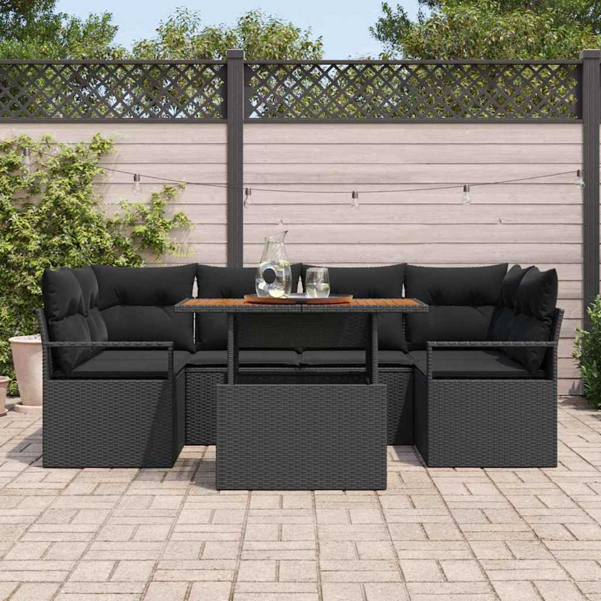 Garten-Set: schwarzes Korbmöbel mit Kissen, Holztisch, Glaskrug und Gläser. Auf einer Ziegelterrasse mit Holzzaun.