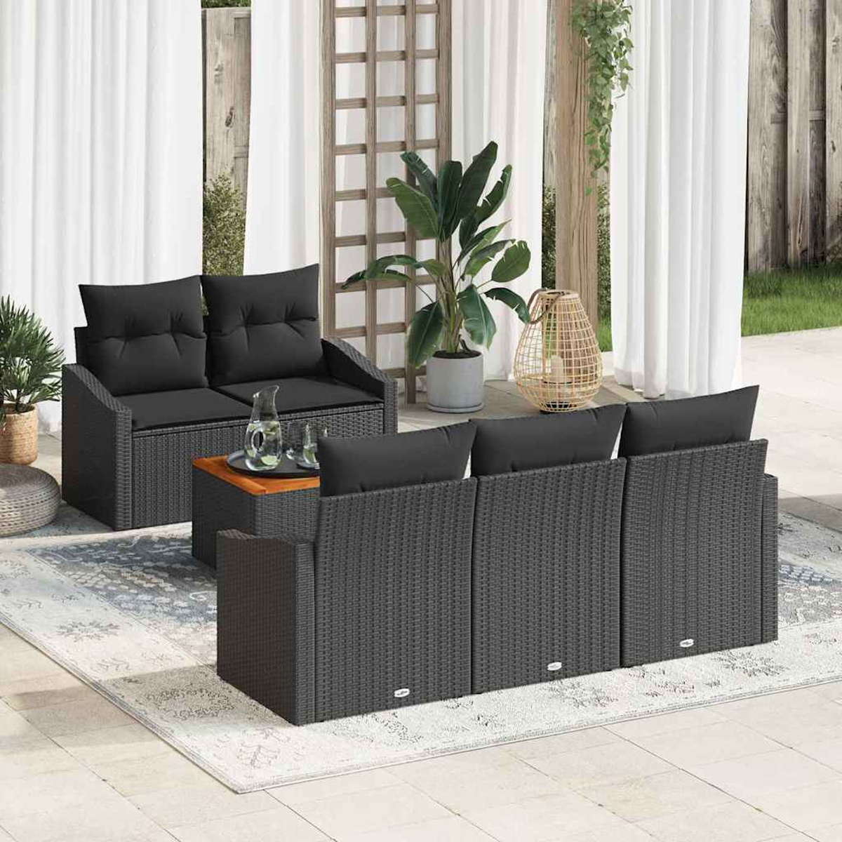 Garten-Set mit grauen Korbmöbeln, schwarzen Kissen, Holztisch und Pflanzen. Weiße Vorhänge und ein Teppich vervollständigen die Szene.