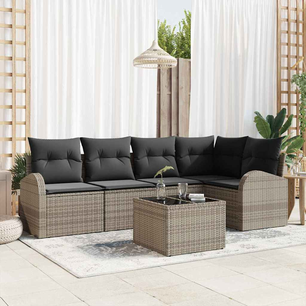 Outdoor-Sitzecke mit grauen Kissen, Rattanmöbeln, einem Couchtisch und weißen Vorhängen. Hintergrund mit Grün.