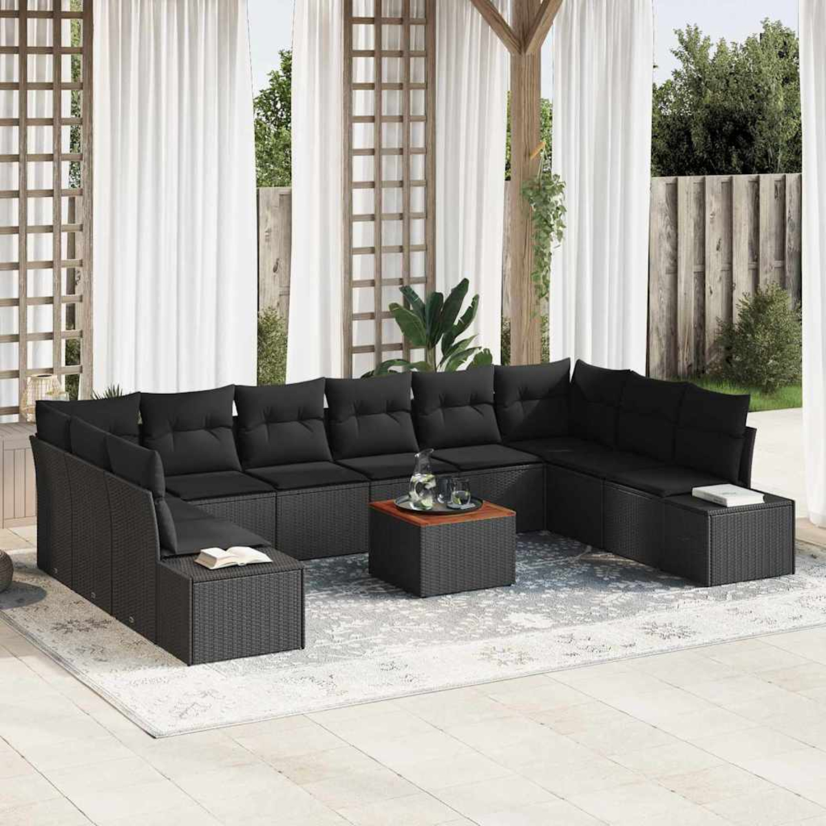 Outdoor-Set mit schwarzem Korbsessel, Kissen und Holztisch. Weiße Vorhänge und Grün im Hintergrund.