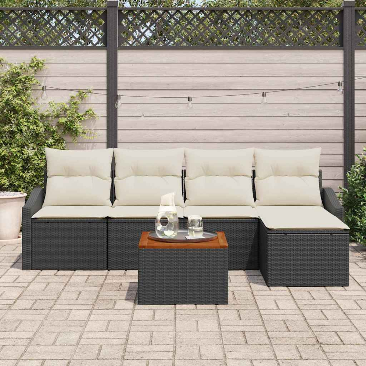 Juego de patio al aire libre con un sofá de mimbre negro y una mesa de café. Cojines beige, jarra de agua y vasos sobre la mesa.