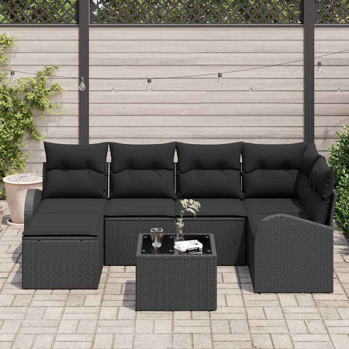 Schwarzes Outdoor-Rattan-Ecksofa mit schwarzen Kissen und einem kleinen Tisch. Im Hintergrund ein Holzzaun und Pflanzen.