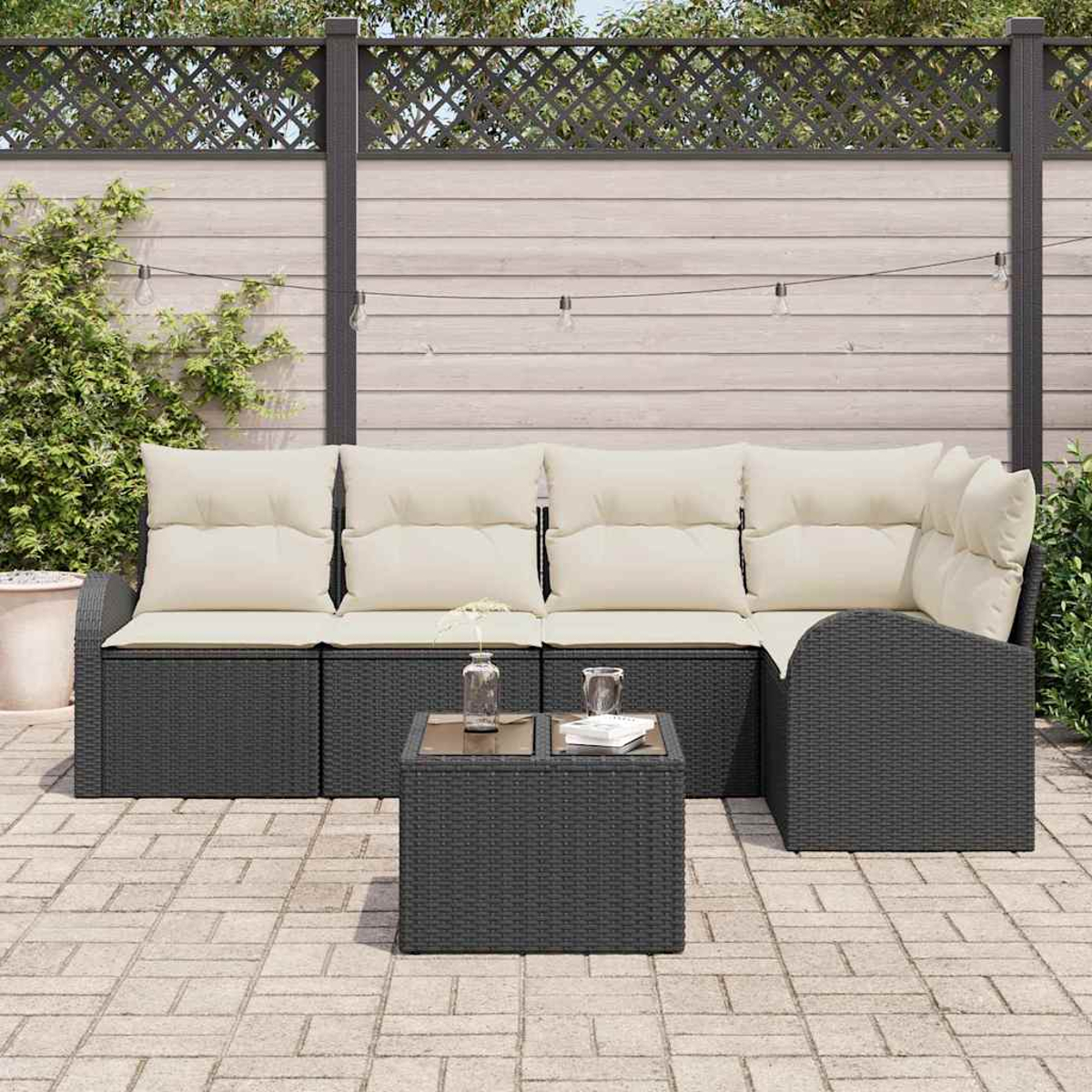 Juego de patio al aire libre con muebles de mimbre grises, cojines crema y una mesita. Una valla y plantas verdes en el fondo.