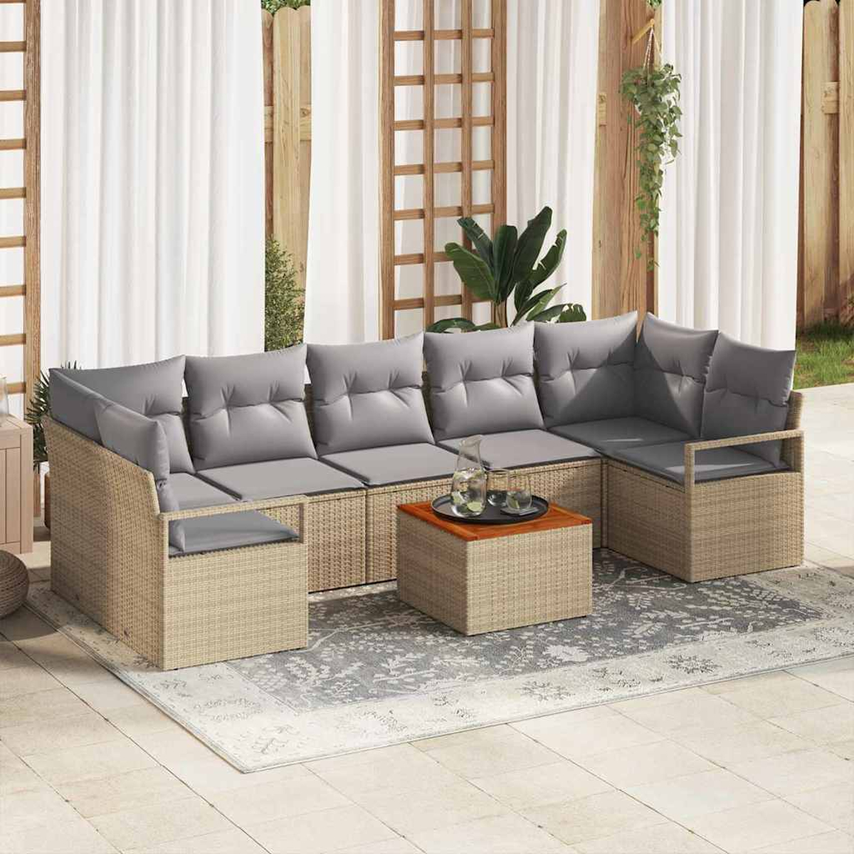 Rattan-Sofa im Freien mit grauen Kissen und einem Holztisch auf einem gemusterten Teppich. Hintergrund: Holzzaun, weiße Vorhänge.