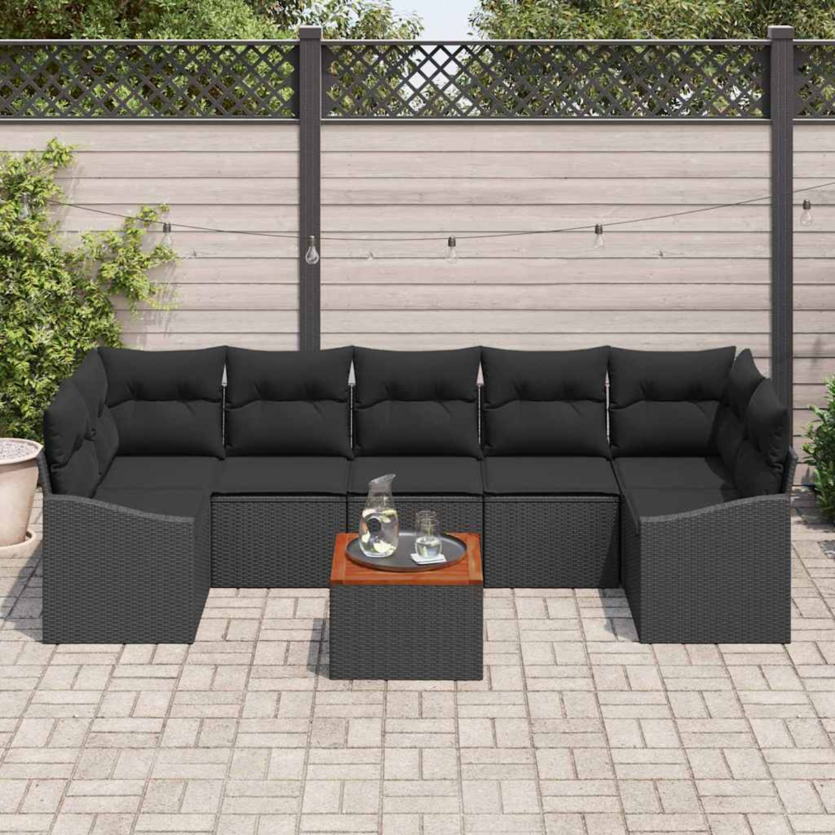 Schwarzes Outdoor-Korbmöbel mit schwarzen Kissen, einem Tisch mit Holzplatte und Wasserkrug auf einer Terrasse.