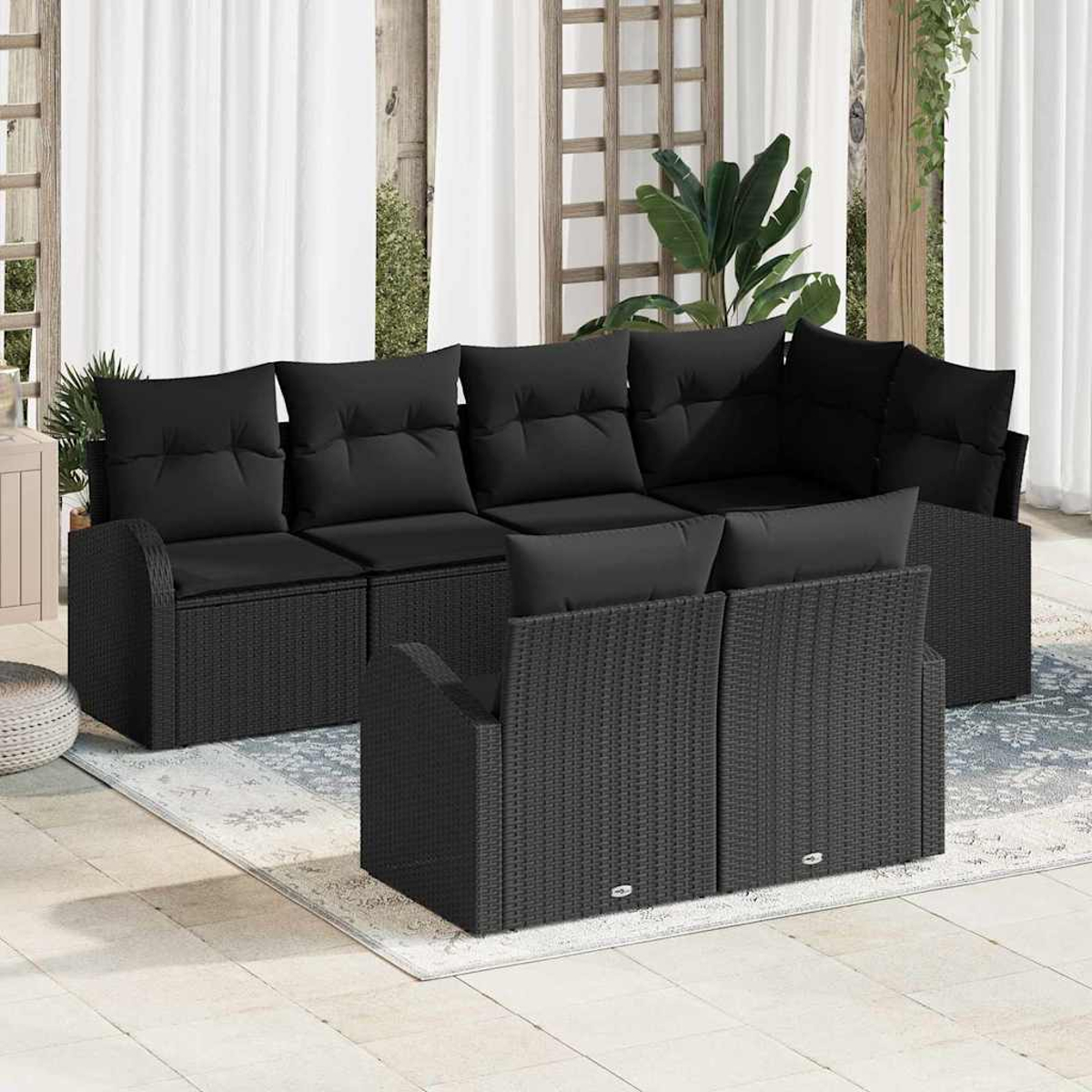 Ein schwarzes Outdoor-Ecksofa aus Korbgeflecht mit Kissen. Es steht auf einem gemusterten Teppich, im Hintergrund weiße Vorhänge.