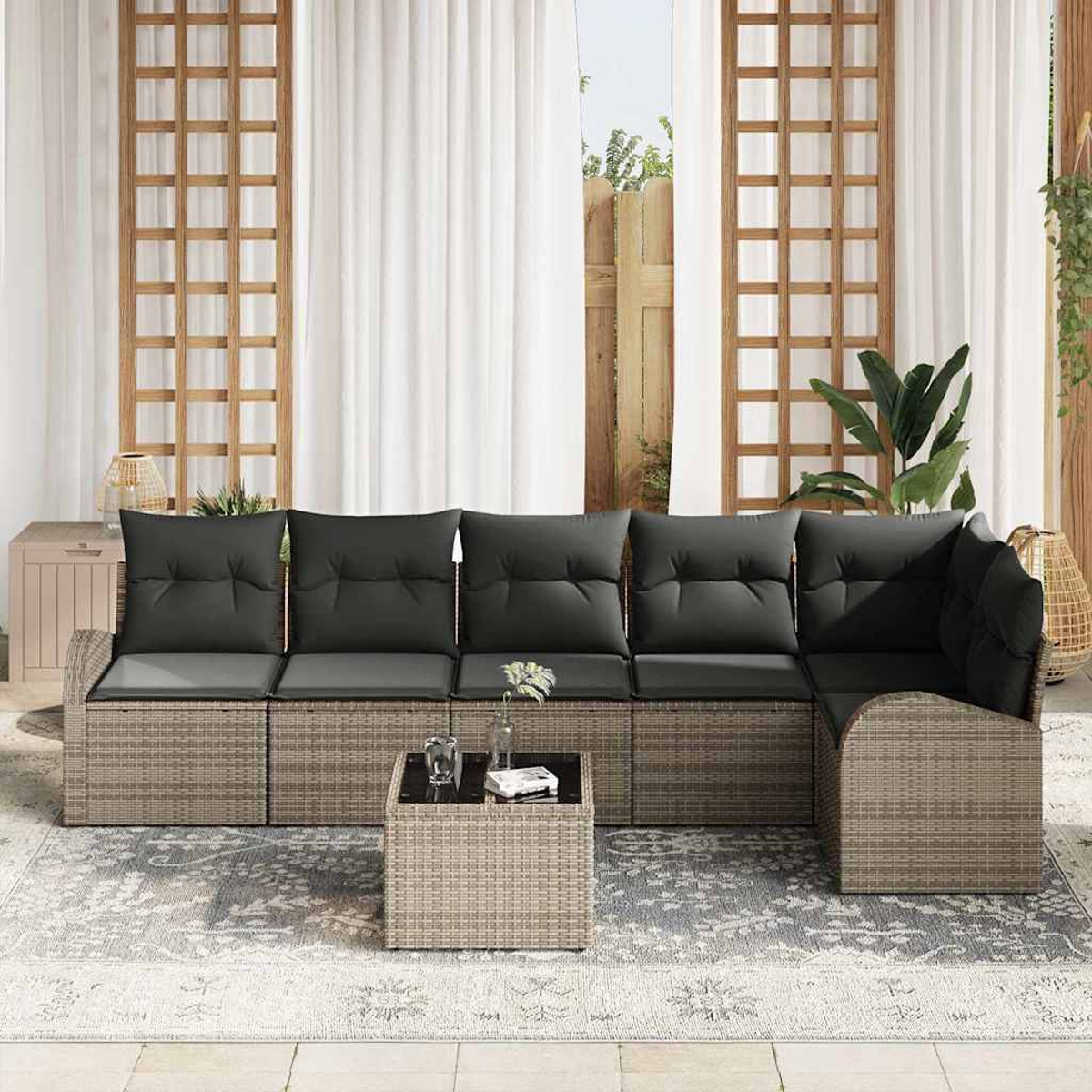 Outdoorsofa mit grauen Kissen und passendem Beistelltisch auf einem gemusterten Teppich. Weiße Vorhänge und Holzgitter dahinter.