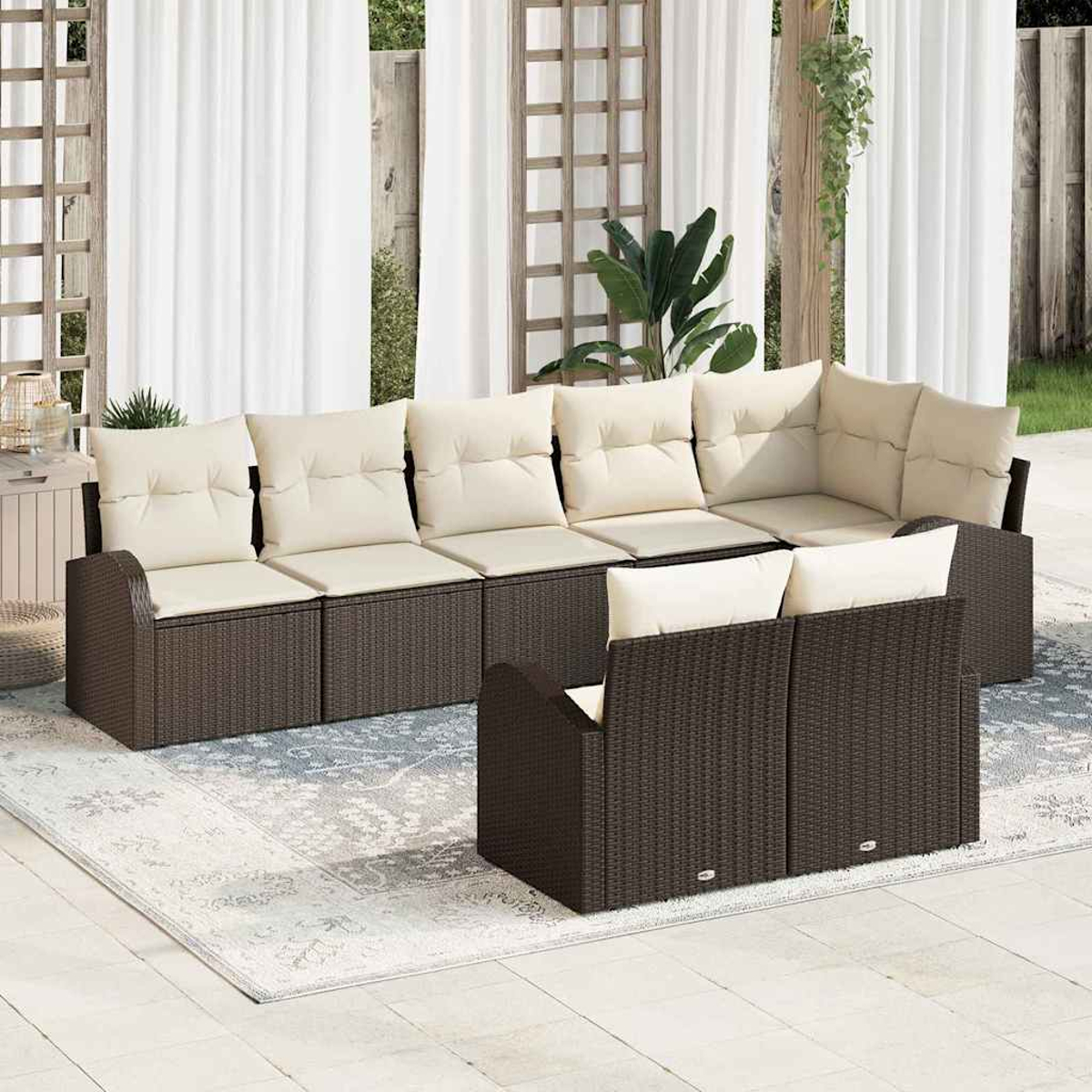 Outdoor-Sitzgruppe mit braunen Korbflechtrahmen und cremefarbenen Kissen. Auf der Terrasse liegt ein Teppich. Weiße Vorhänge und Grünzeug im Hintergrund.