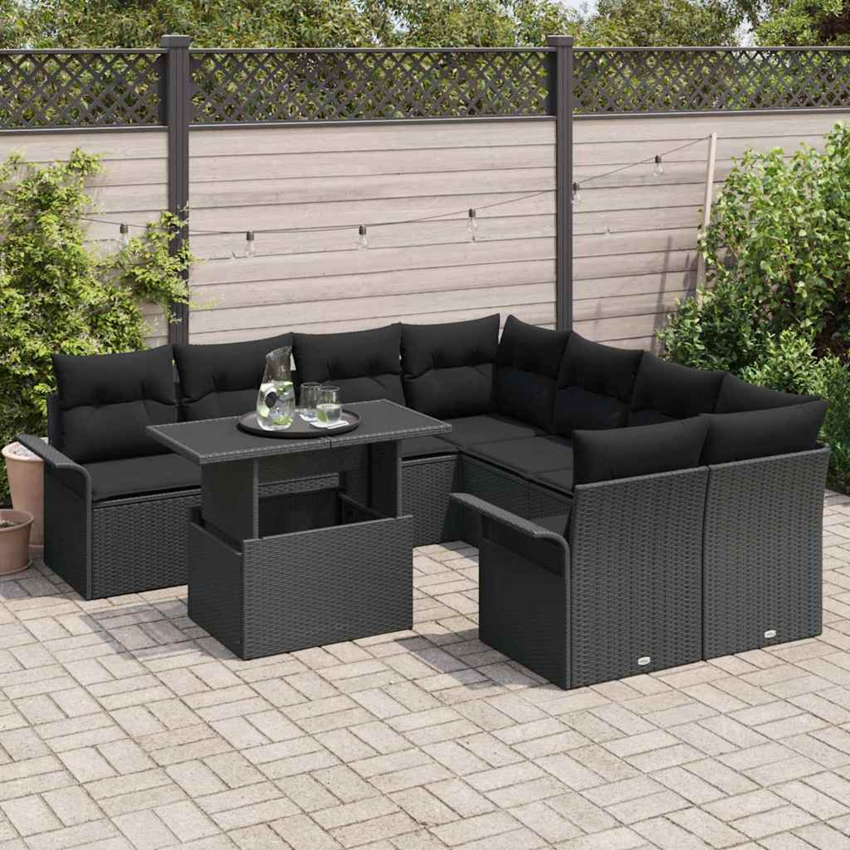 Garten-Sitzgruppe mit schwarzen Kissen und einem Tisch mit Hubfunktion auf einer gepflasterten Terrasse. Im Hintergrund ein Zaun und Grün.