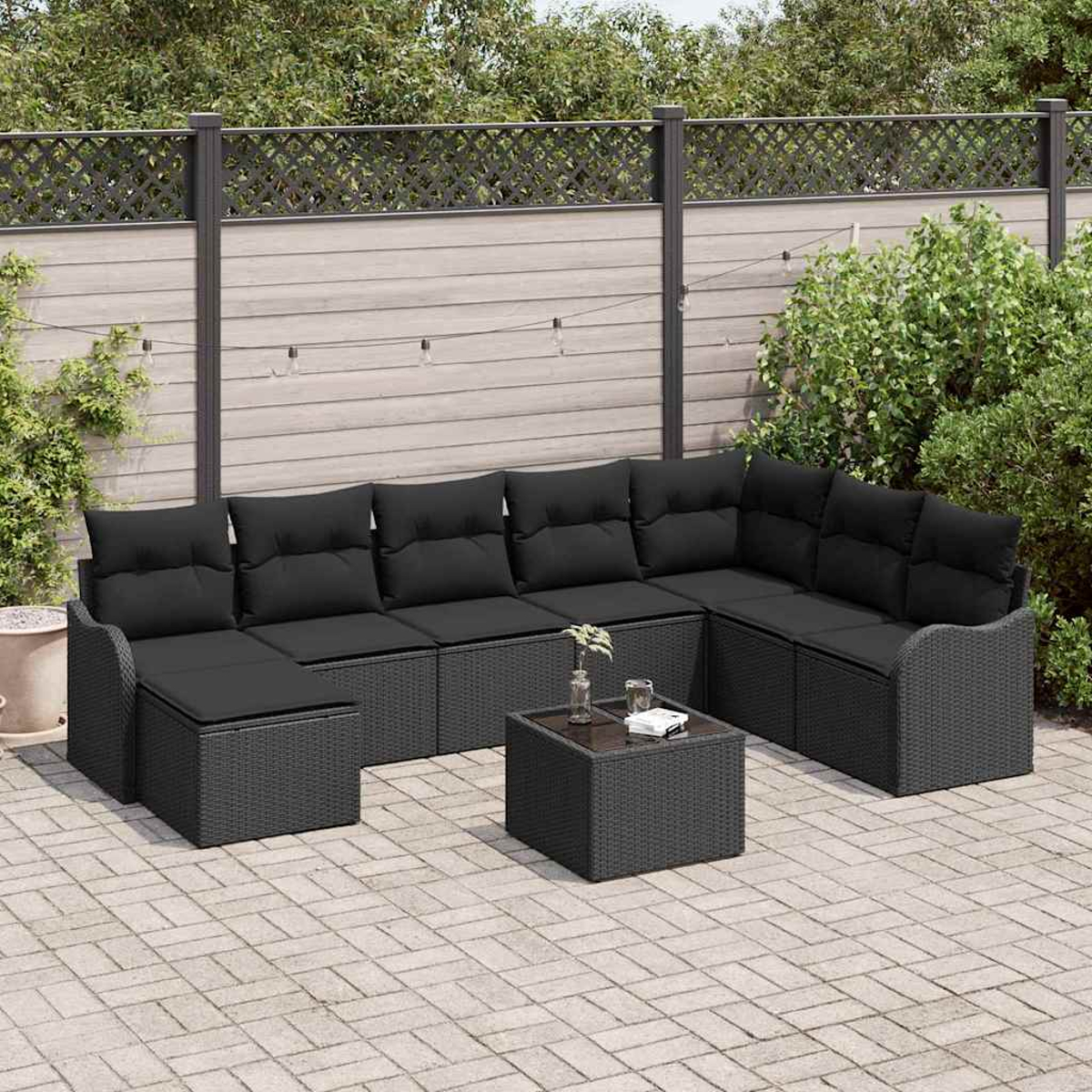 Gartenmöbel-Set: Schwarzes Ecksofa und quadratischer Tisch auf gepflasterter Terrasse. Grüne Pflanzen und Holzzaun im Hintergrund.
