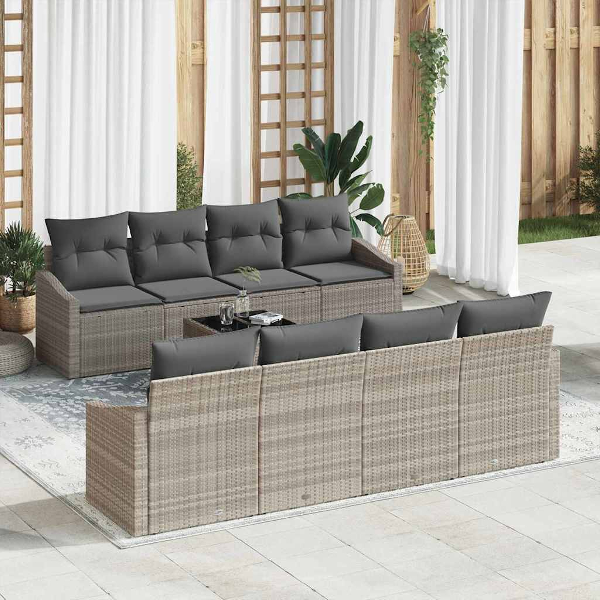 Terrassengarnitur mit grauen Kissen. Korbsessel, Couchtisch und weiße Vorhänge im Hintergrund. Pflanzen und ein Teppich sind sichtbar.