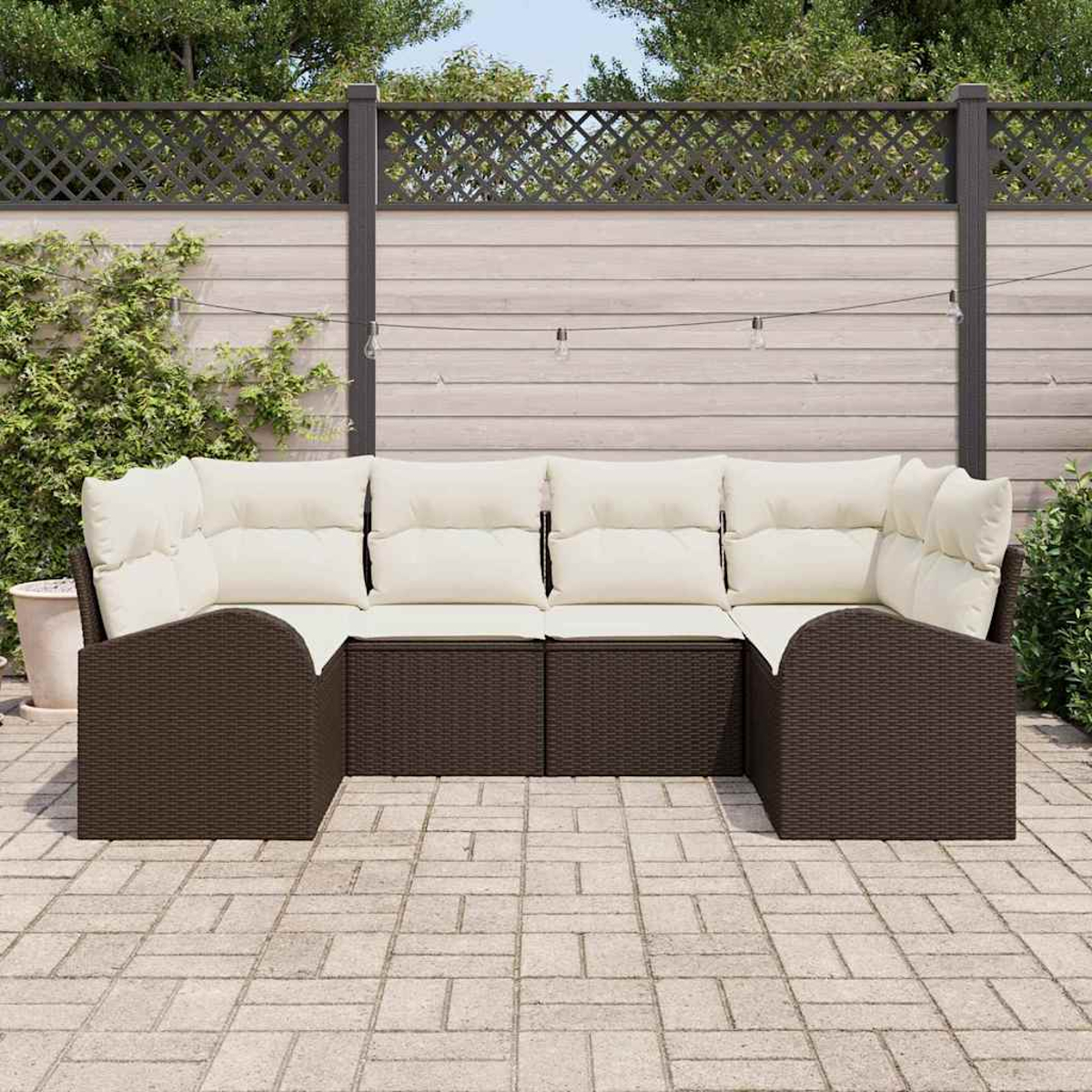 Ein braunes Korbsessel-Ecksofa mit cremefarbenen Kissen steht auf einer gepflasterten Terrasse. Im Hintergrund befinden sich ein Holzzaun und Grün.