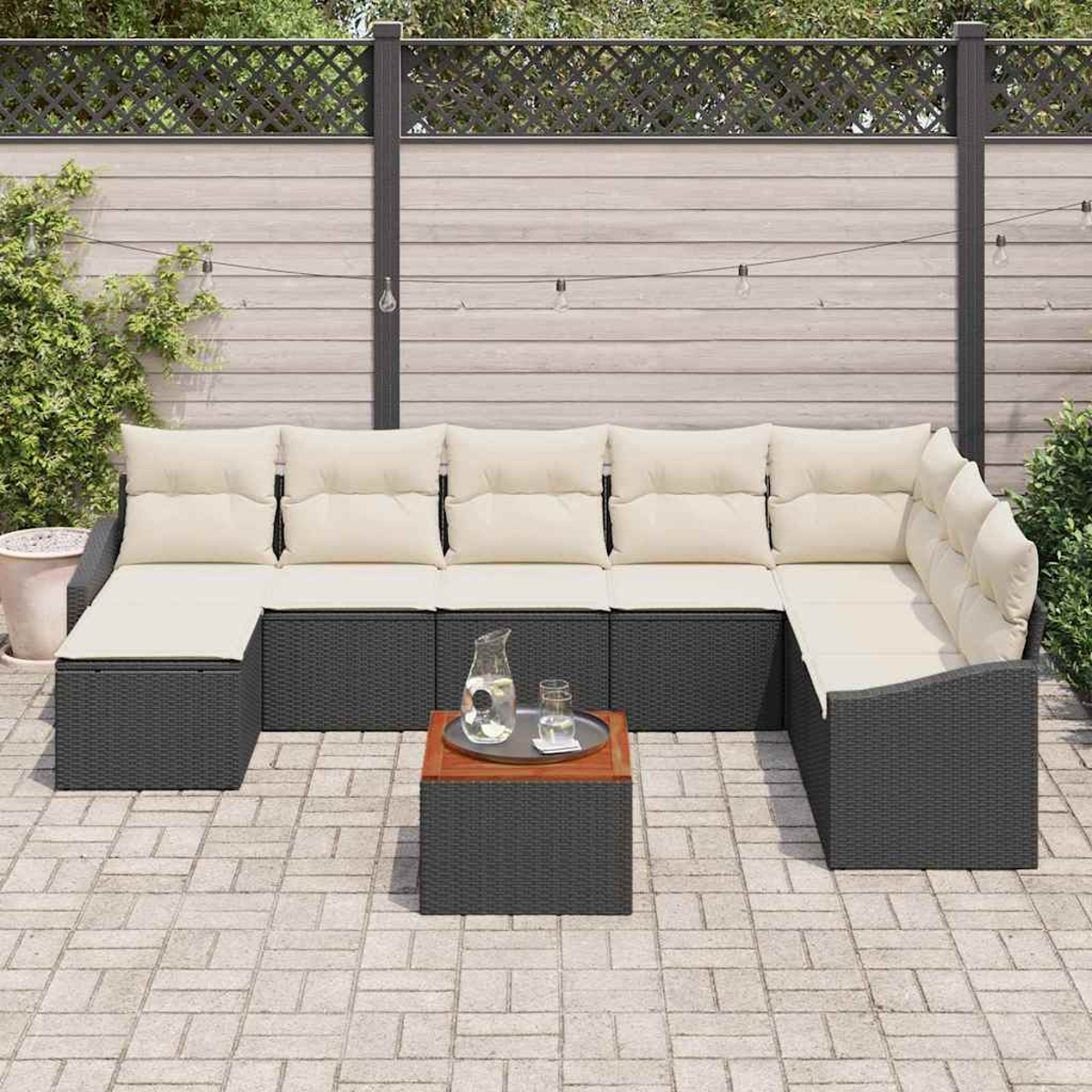 Outdoor-Ecksofa mit cremefarbenen Kissen und einem kleinen Tisch mit Glaskrug und Tablett. Zaun und Grün im Hintergrund.