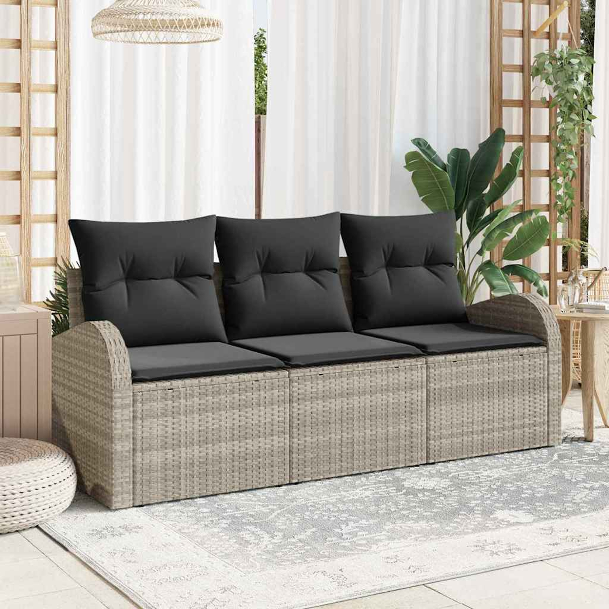 Ein graues Rattansofa mit schwarzen Kissen steht auf einem Teppich. Dahinter ein Fenster mit weißen Vorhängen und ein Holzgitter mit Pflanzen.