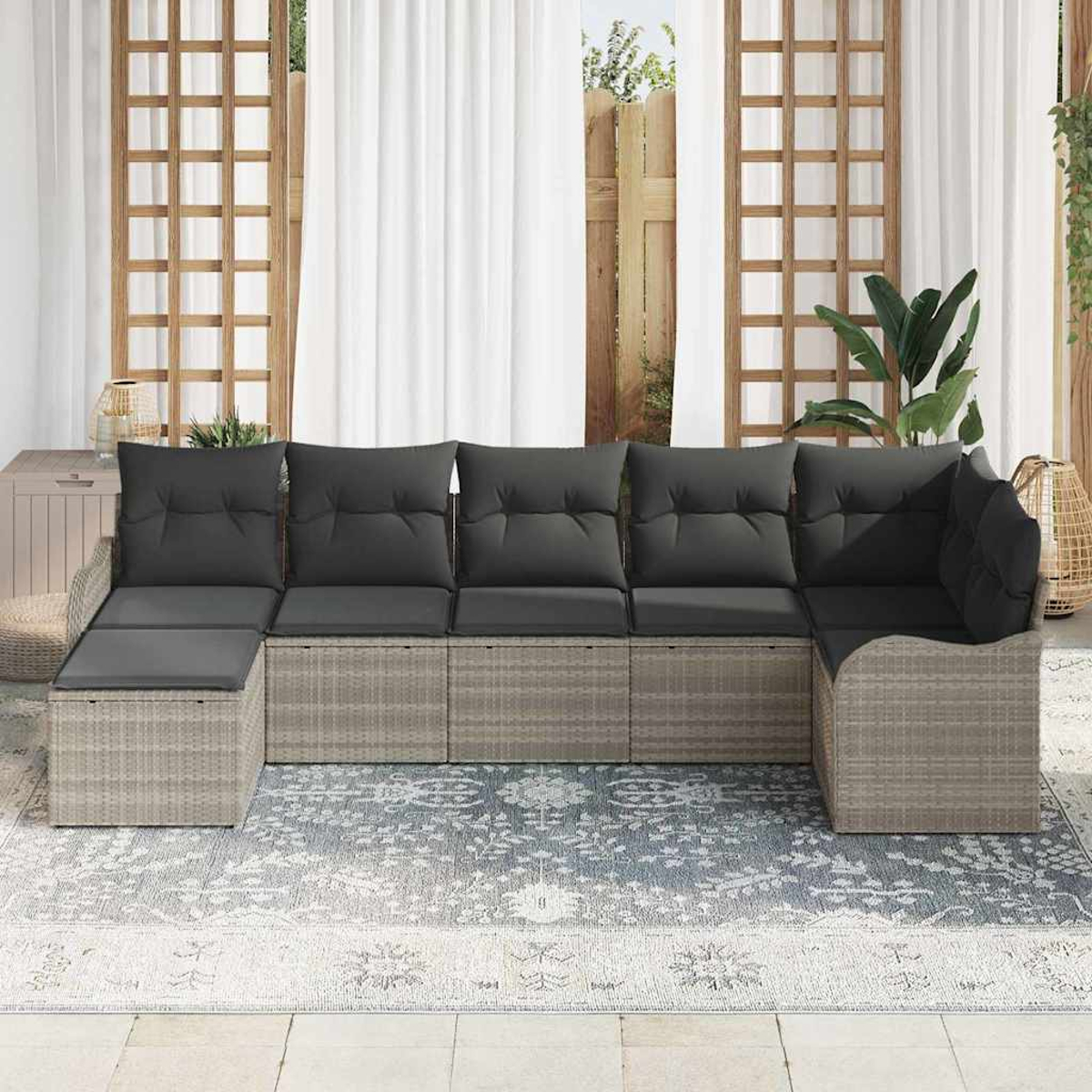 Ein Ecksofa im Freien mit grauen Kissen steht auf einem gemusterten Teppich. Weiße Vorhänge und ein Holzgitter sind im Hintergrund.