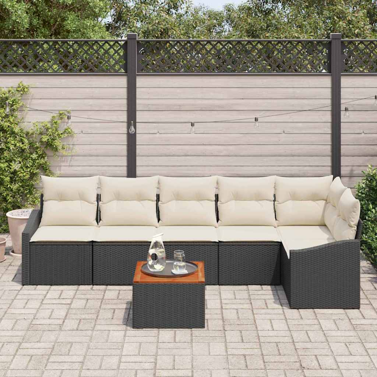 Terrassenset mit Ecksofa, beigen Kissen und kleinem Tisch mit Tablett. Im Hintergrund ein Holzzaun und Grün.