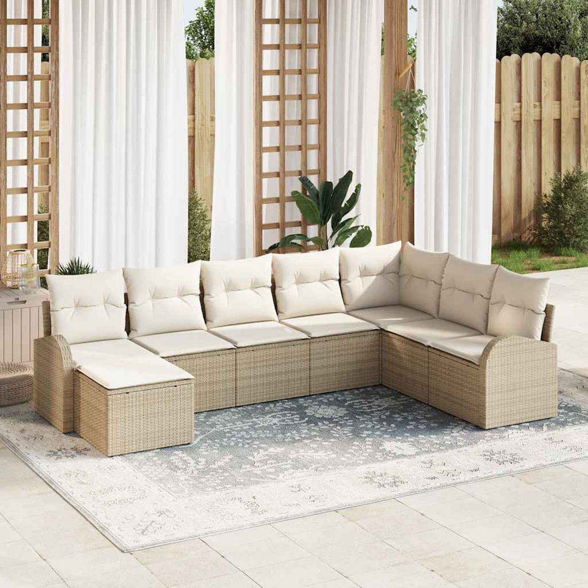 Un sofá seccional al aire libre con cojines beige y base tejida se encuentra sobre una alfombra estampada. Cortinas blancas y una valla de madera al fondo.