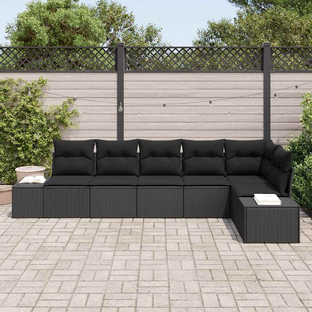 Schwarzes Rattan-Ecksofa im Freien mit schwarzen Kissen auf einer gefliesten Terrasse. Ein Zaun und Grün im Hintergrund.
