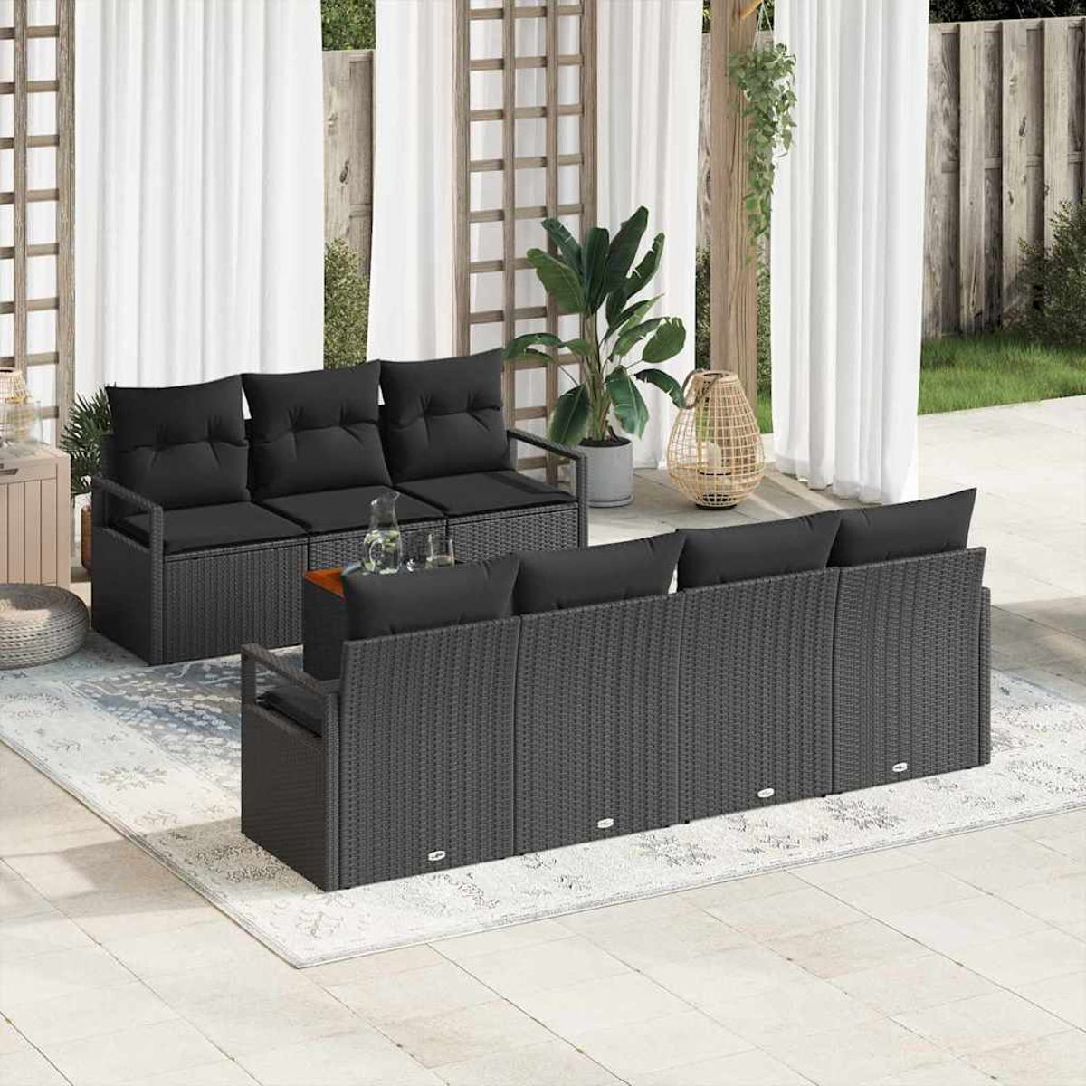 Außenbereich mit dunkelgrauen Rattan-Sofas und schwarzen Kissen, einem Teppich und Pflanzen. Weiße Vorhänge und Holzgitter im Hintergrund.