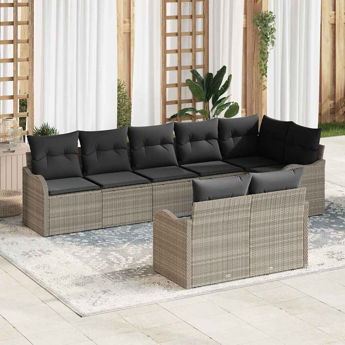 Rattan-Sofa im Freien mit schwarzen Kissen. Weiße Vorhänge und Holzpaneele im Hintergrund. Ein Teppich bedeckt den Boden.