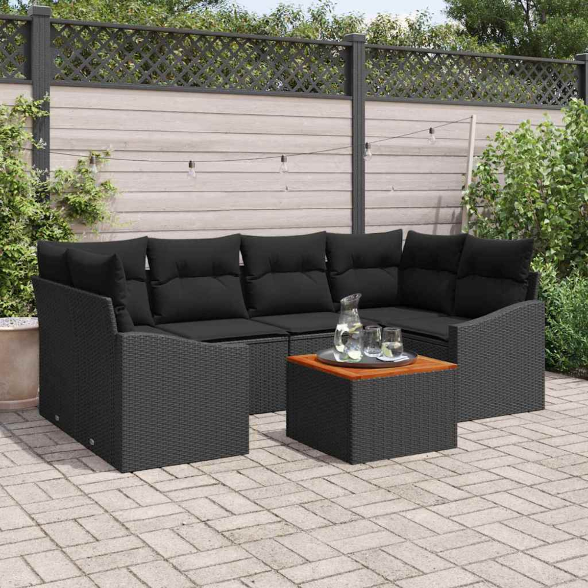Garten-Sitzgruppe mit schwarzen Kissen. Ein kleiner Tisch mit Holzplatte hält Gläser und einen Krug. Hintergrund: Zaun, Grün.
