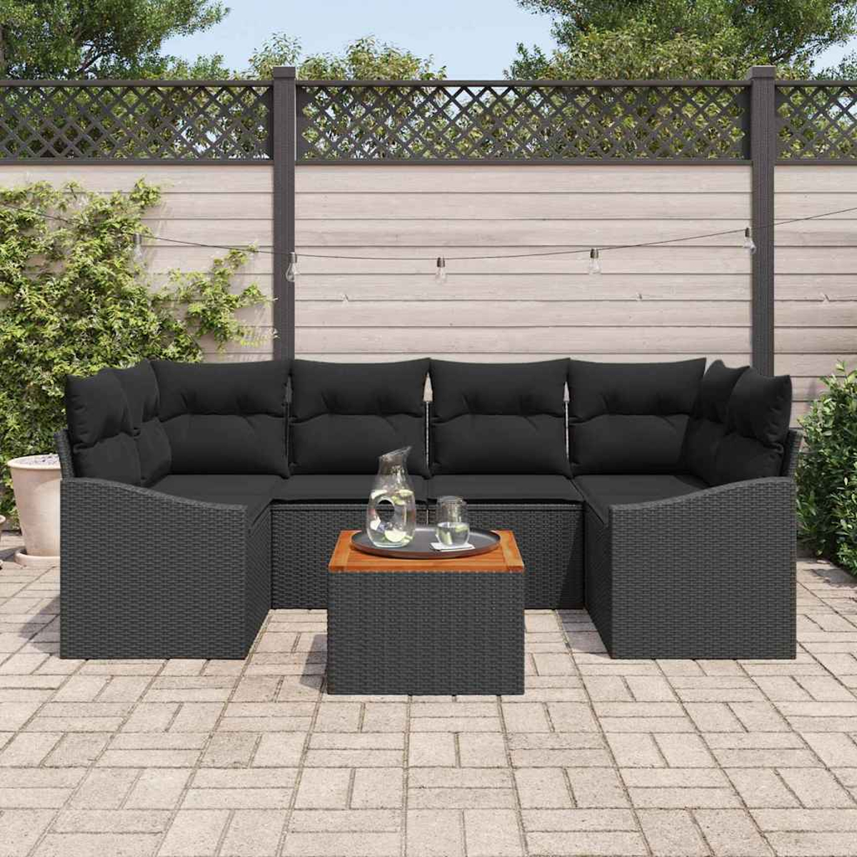 Garten-Sitzgruppe mit schwarzen Kissen und grauem Korbgewebe. Ein Holztisch trägt Krug und Glas. Hintergrund ist ein Holzzaun.