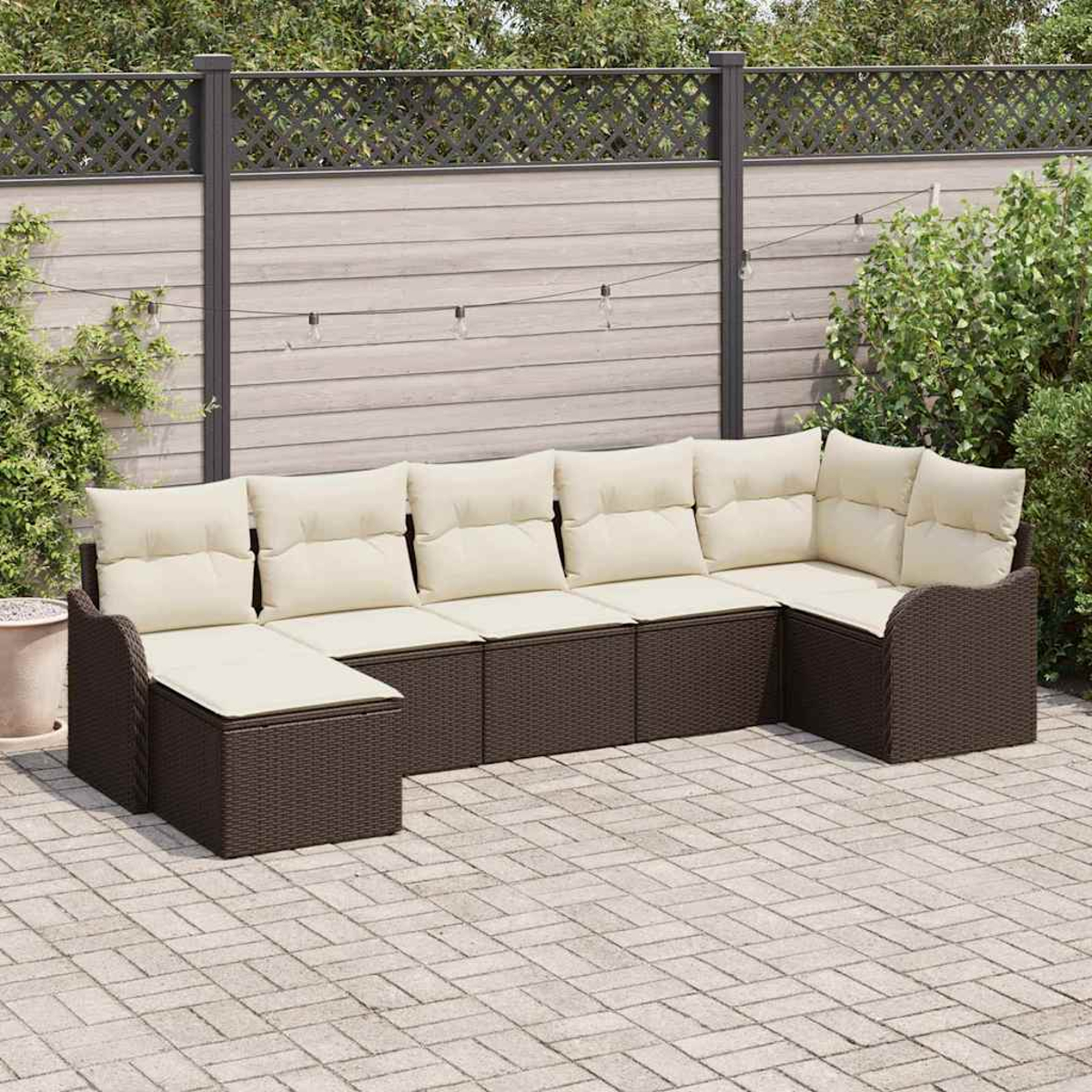 Ein braunes Korbsessel-Sofa mit cremefarbenen Kissen steht auf einer Backsteinterrasse. Ein Holzzaun und Grün bilden den Hintergrund.
