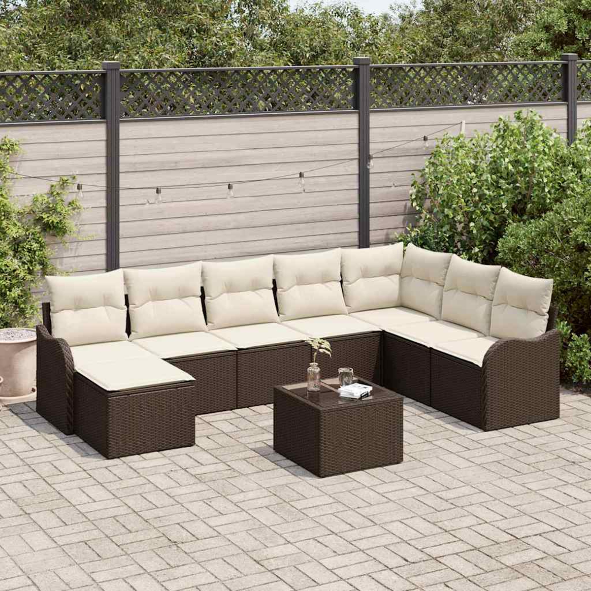 Outdoor-Ecksofa mit cremefarbenen Kissen und dunkelbraunem Korbgeflecht. Davor steht ein kleiner quadratischer Tisch. Im Hintergrund ein Zaun und Grün.