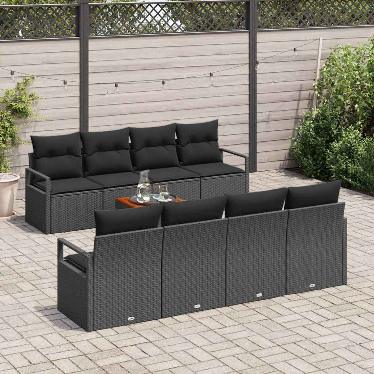 Szene im Freien mit dunkelgrauen Korbsesseln und schwarzen Kissen auf einer Backsteinterrasse. Ein kleiner Tisch mit Getränken steht zwischen den Sofas.