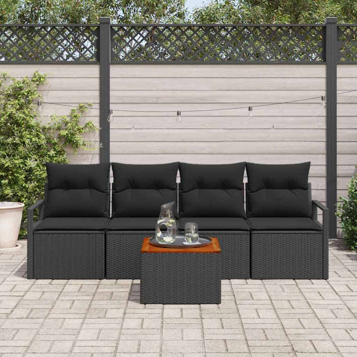 Gartenmöbel: ein dunkelgraues, gewebtes Sofa mit schwarzen Kissen, ein kleiner Tisch mit Glaskrug und Gläsern. Hintergrund: ein Holzzaun.