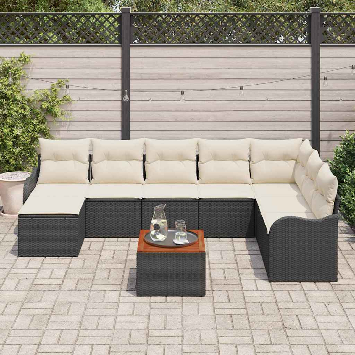 Ein Ecksofa mit cremefarbenen Kissen steht auf einer Terrasse. Ein kleiner Tisch trägt Getränke. Im Hintergrund ein Zaun und Pflanzen.
