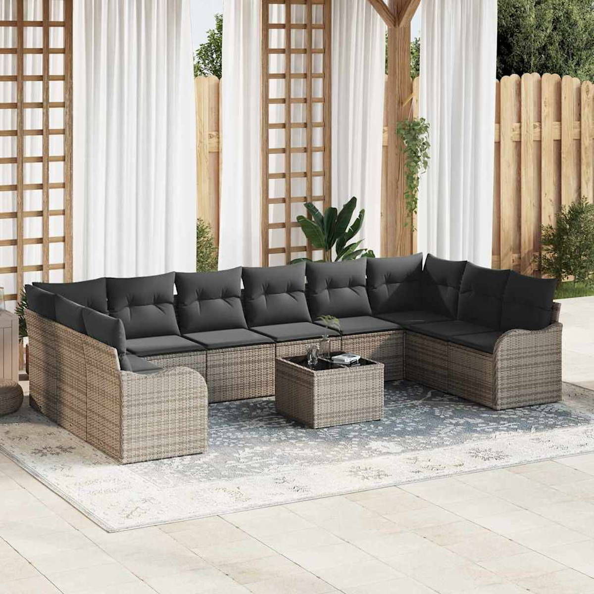 Outdoor-Ecksofa mit grauen Kissen auf einem gemusterten Teppich. Weiße Vorhänge, Holzzaun und Grünzeug vervollständigen die Szene.