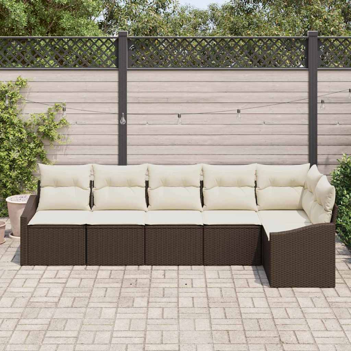Outdoor-Ecksofa mit cremefarbenen Kissen, braunem Korbgewebe, auf einer Backsteinterrasse. Dahinter ein Holzzaun und Grün.