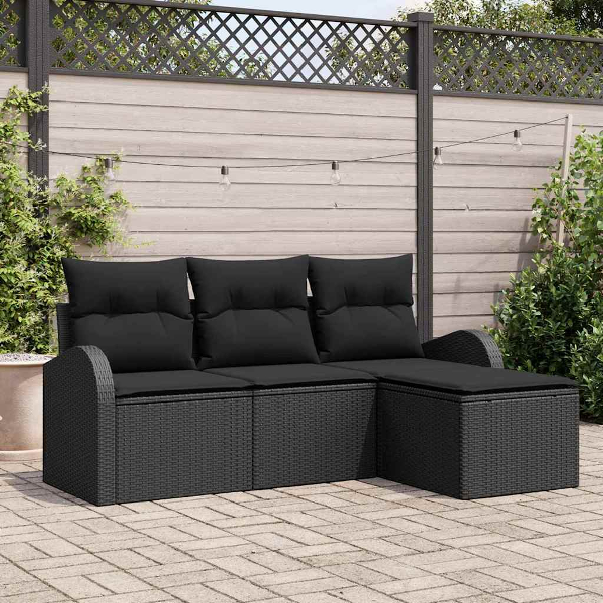 Ein schwarzes Korbmöbel mit schwarzen Kissen steht auf einer Terrasse. Ein Zaun und Pflanzen sind im Hintergrund.