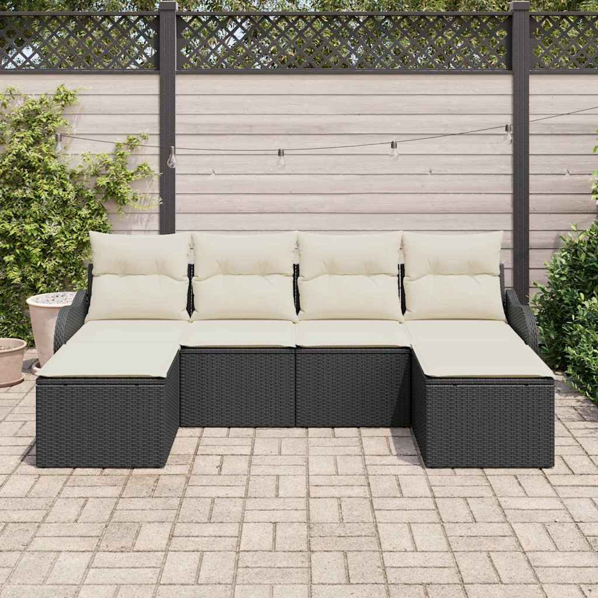 Ein schwarzes Korbsessel-Sofa mit cremefarbenen Kissen steht auf einer Terrasse. Ein Holzzaun und Pflanzen sind im Hintergrund.