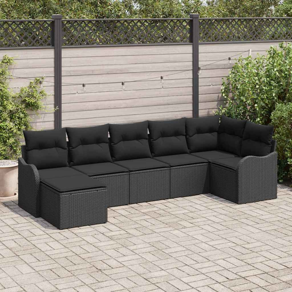 Ein schwarzes Rattan-Ecksofa mit schwarzen Kissen steht auf einer Backsteinterrasse. Im Hintergrund befinden sich ein Holzzaun und Grün.