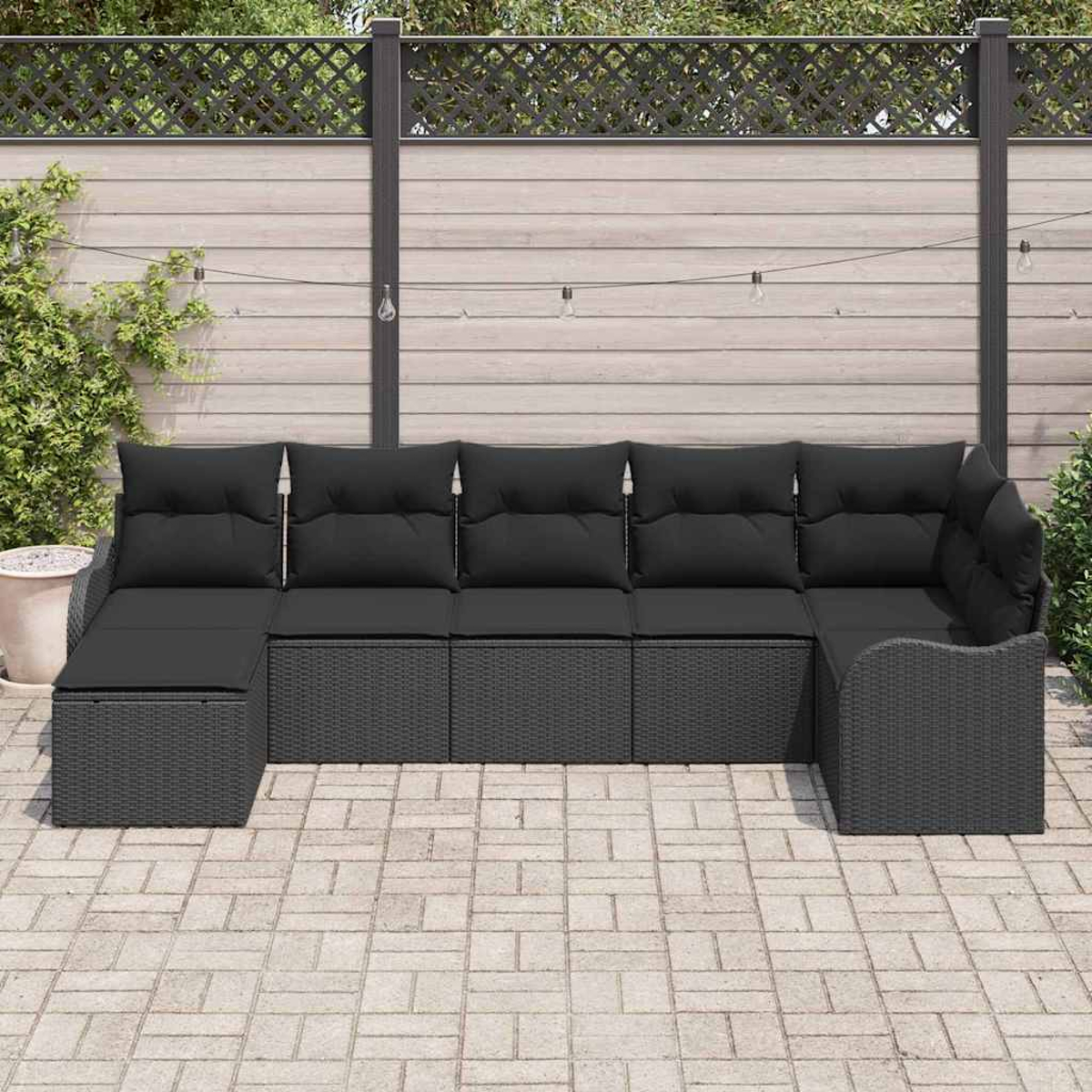 Ein schwarzes Rattan-Ecksofa steht auf einer Terrasse. Es hat schwarze Kissen und ist L-förmig angeordnet. Dahinter ist ein Zaun.