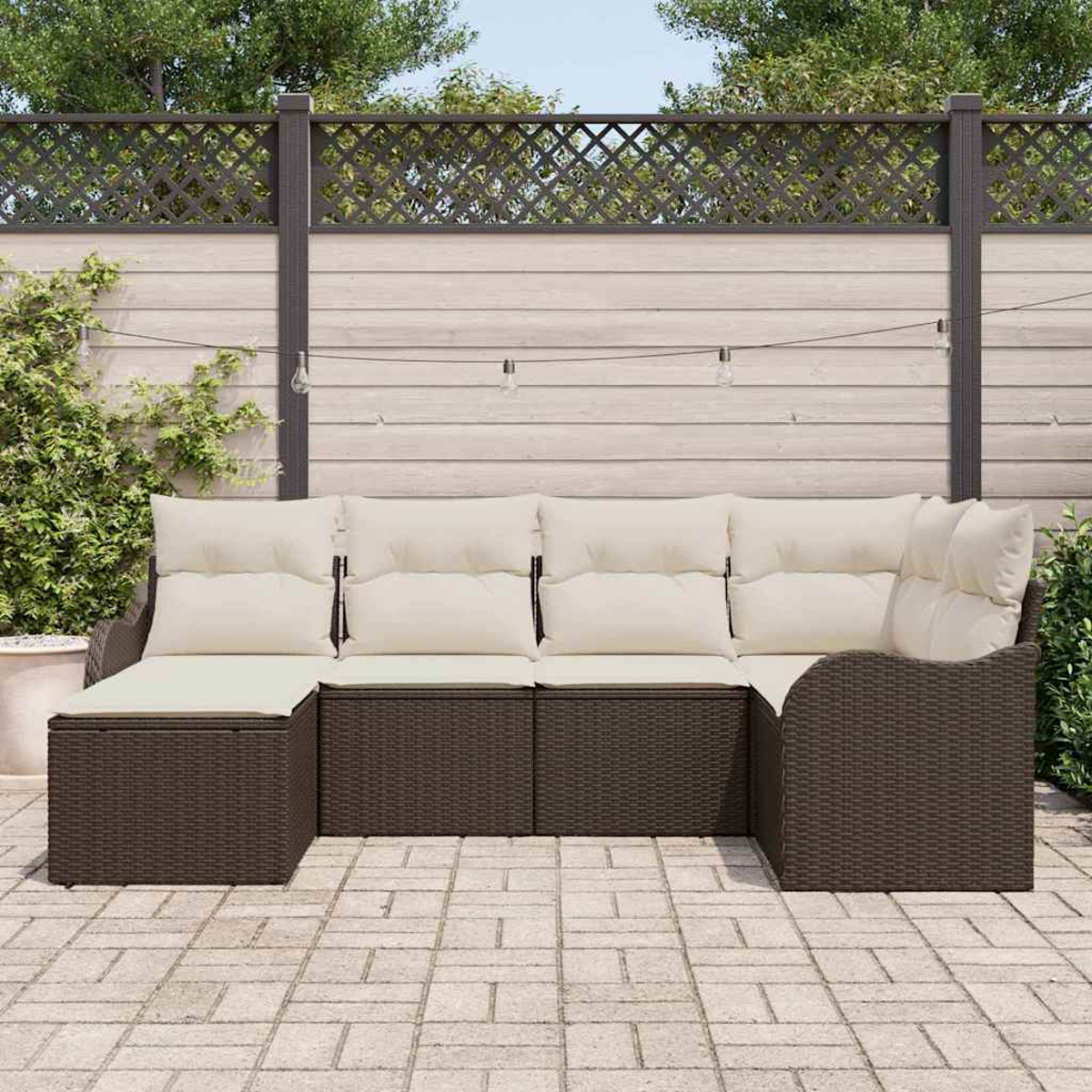Ein braunes Korbsessel-Sofa mit cremefarbenen Kissen steht auf einer Steinterrasse. Im Hintergrund befinden sich ein Holzzaun und Grün.