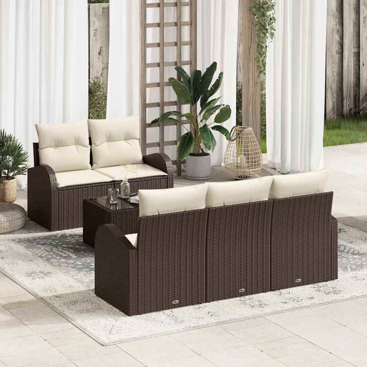 Terrassenmöbel-Set mit braunen Korbmöbeln, cremefarbenen Kissen, Glastisch und Pflanzen in einem Garten.