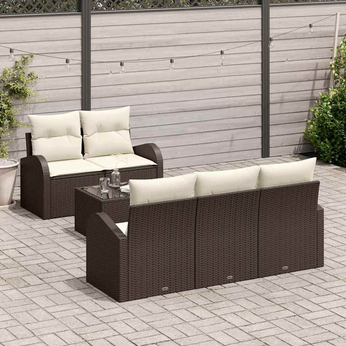 Gartenmöbel-Set. Braunes Korbmöbel-Sofa und -Liebessessel mit cremefarbenen Kissen. Tisch mit Glasplatte. Lichterkette und Holzzaun im Hintergrund.