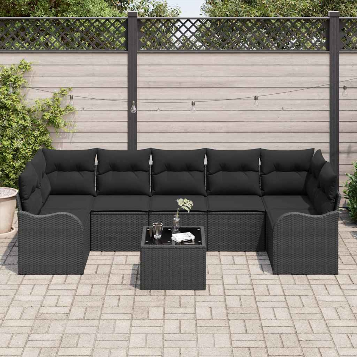 Schwarzes Outdoor-Rattan-Ecksofa mit schwarzen Kissen und einem kleinen Tisch auf einer Backstein-Terrasse, vor einem Holzzaun.