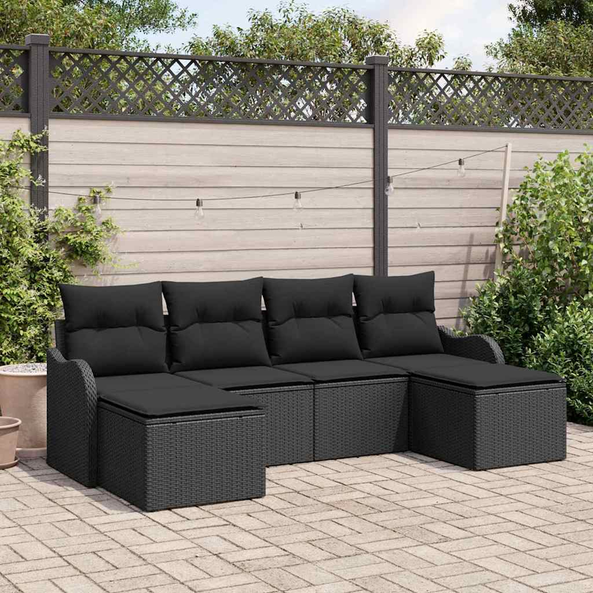 Schwarzes Rattan-Outdoor-Ecksofa auf einer Backstein-Terrasse. Hintergrund: Holzzaun, Lichterketten und Grün.