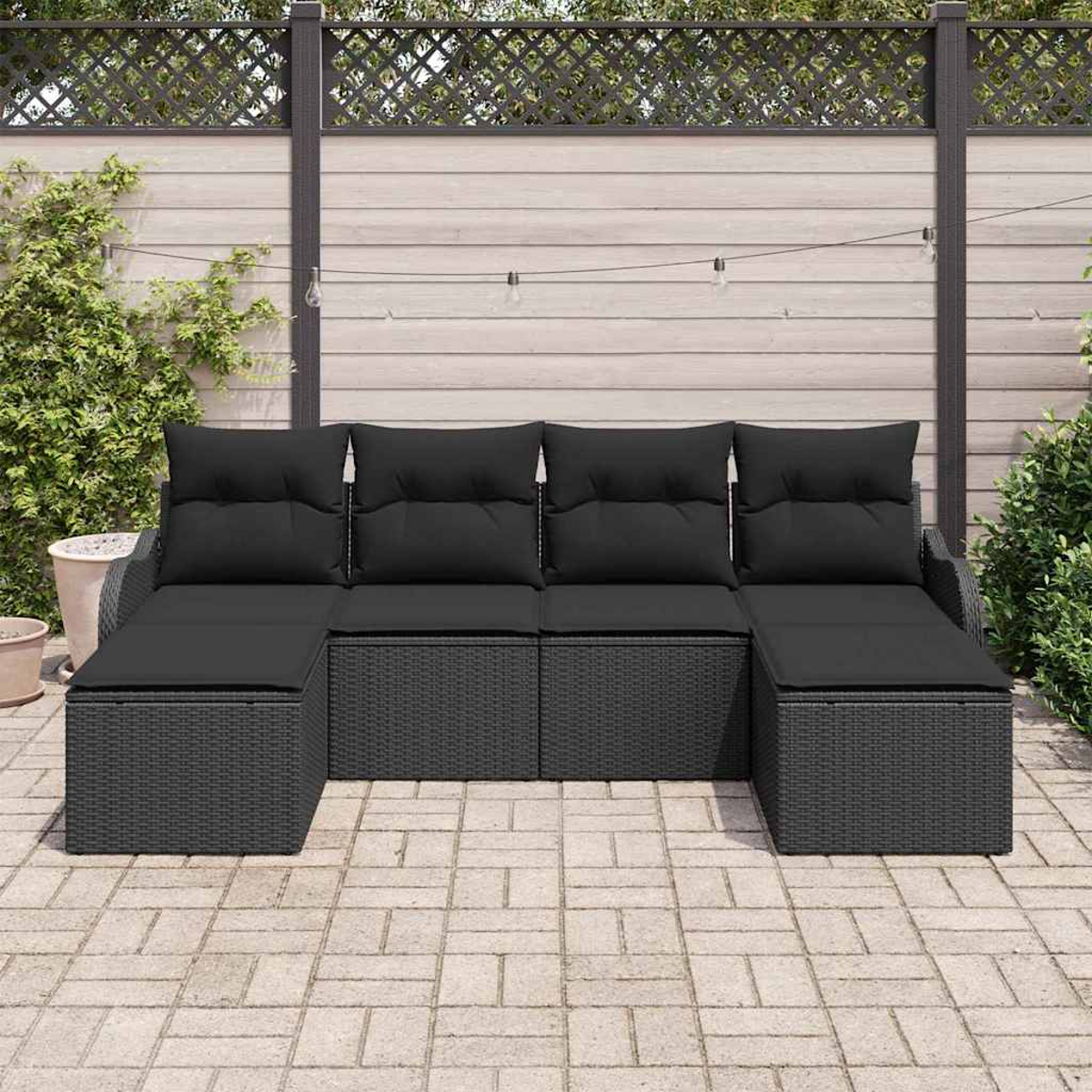 Ein schwarzes Korbmöbel mit schwarzen Kissen auf einer Terrasse. Im Hintergrund ein Holzzaun und Pflanzen. Lichterketten sind sichtbar.
