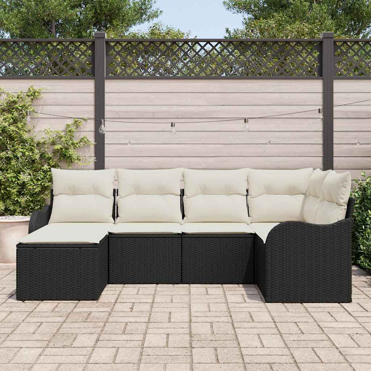 Schwarzes Korbmöbel-Ecksofa mit cremefarbenen Kissen auf einer Backstein-Terrasse, vor einem Holzzaun.