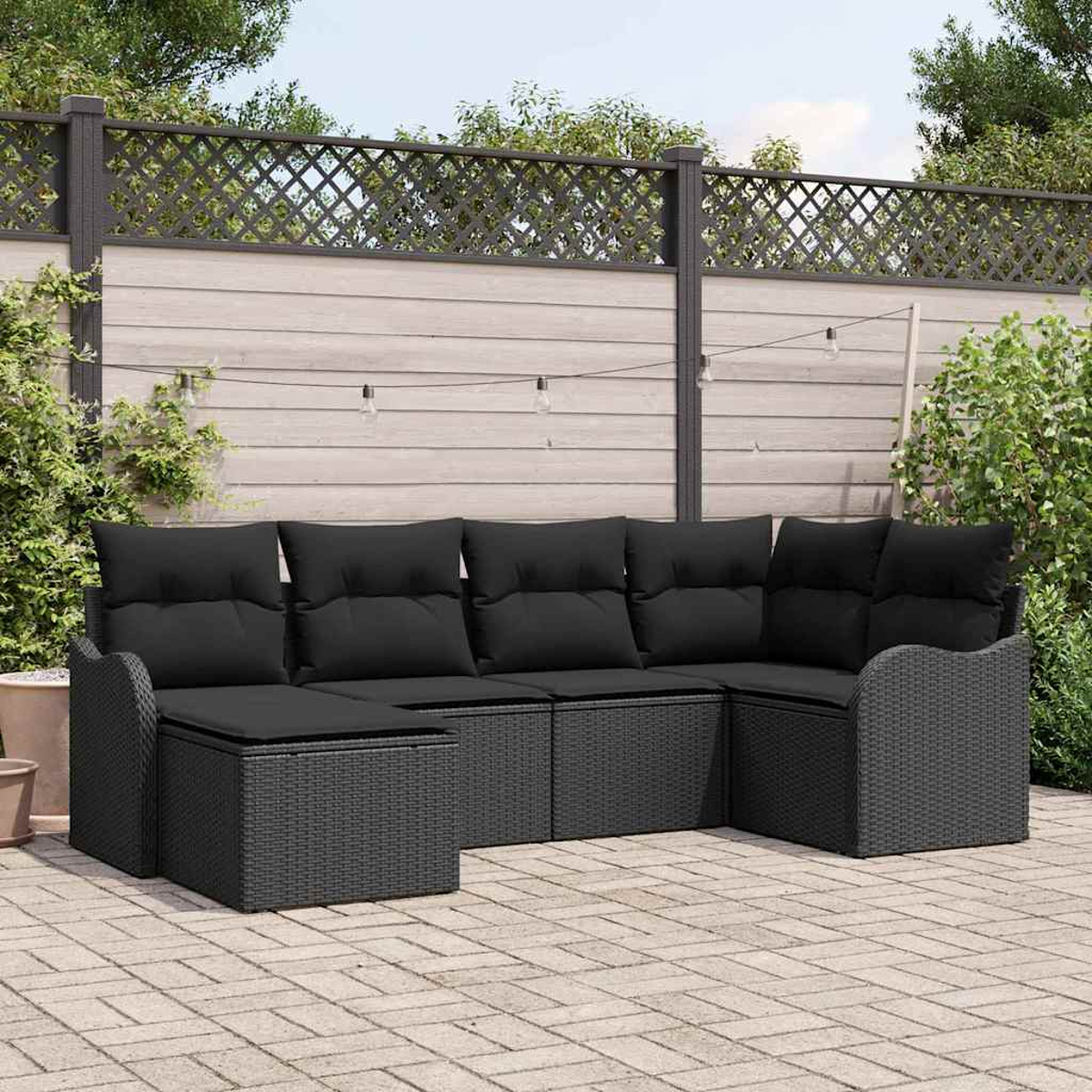 Schwarzes Outdoor-Ecksofa aus Korbgeflecht auf einer Backstein-Terrasse. Im Hintergrund ein Holzzaun, Lichterketten und Grün.