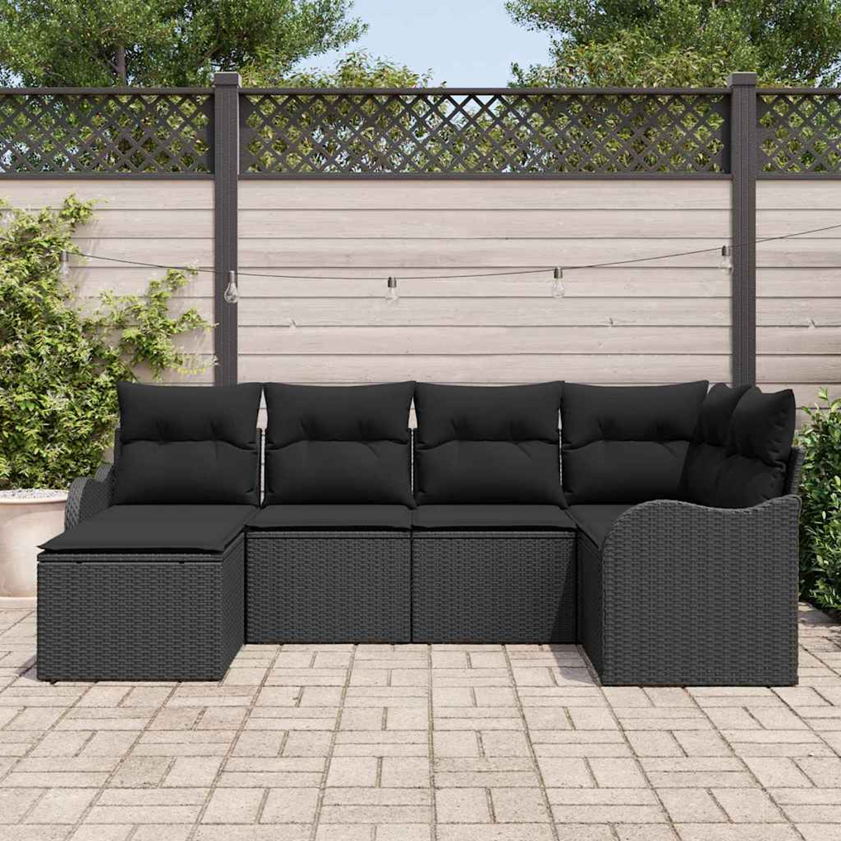 Ein schwarzes Outdoor-Ecksofa aus Korbgeflecht mit schwarzen Kissen steht auf einer Ziegelterrasse. Im Hintergrund ist ein Holzzaun mit Gitteraufsatz.